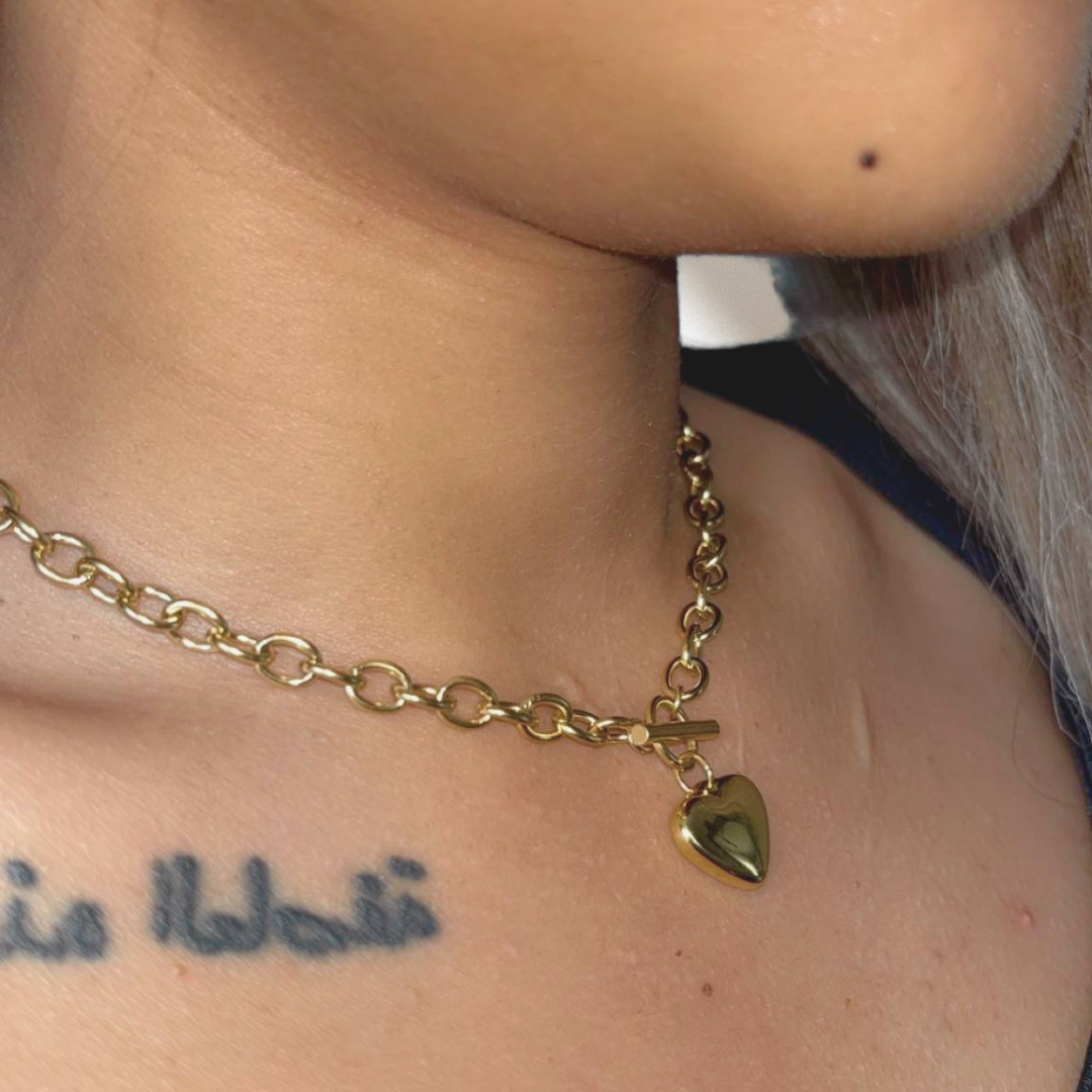 Gold chain necklace with a heart pendant worn by a person with visible tattoos.