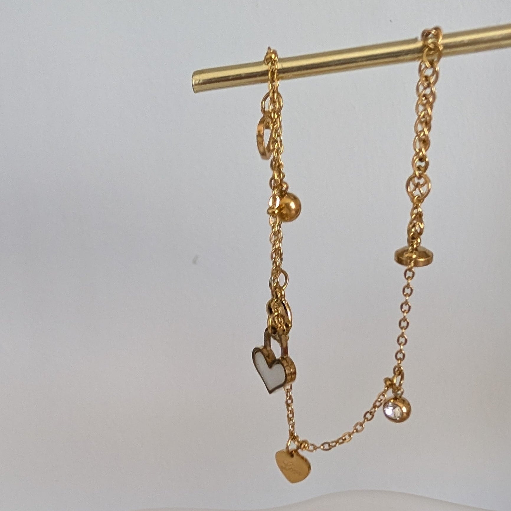 Gold chain bracelet with heart charm on a gold stand against a white background