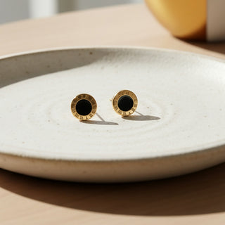 Gold and black stud earrings on a white ceramic plate.