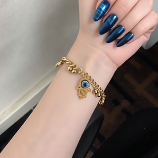 Gold bracelet with a hamsa hand and eye charm on a wrist with blue nail polish.