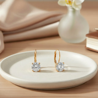 Gold earrings with clear gemstones on a white plate, with a blurred background of a vase and fabric.