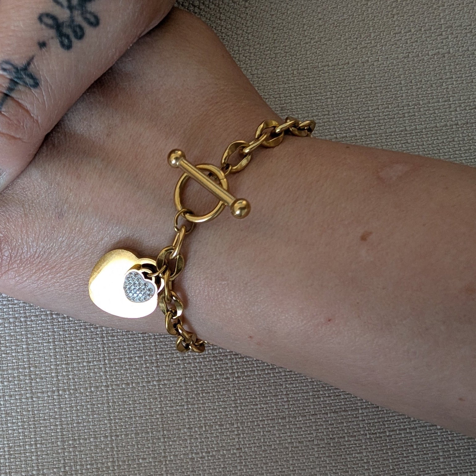Gold bracelet with a heart-shaped charm on a wrist against a neutral background