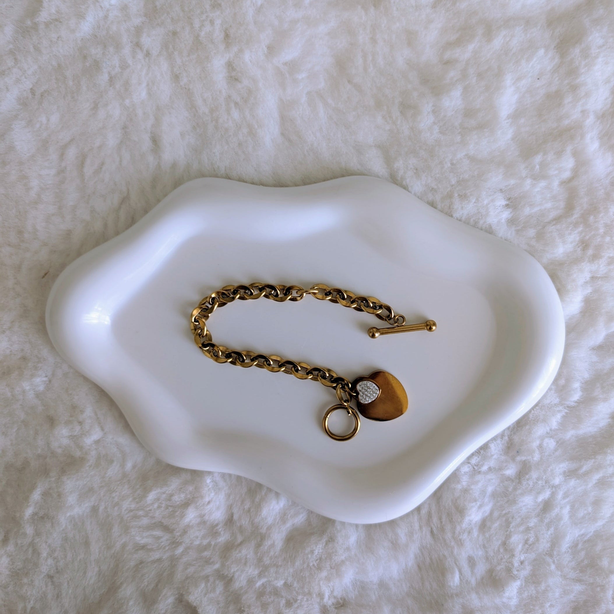 Gold bracelet with a heart charm on a white dish against a textured white background