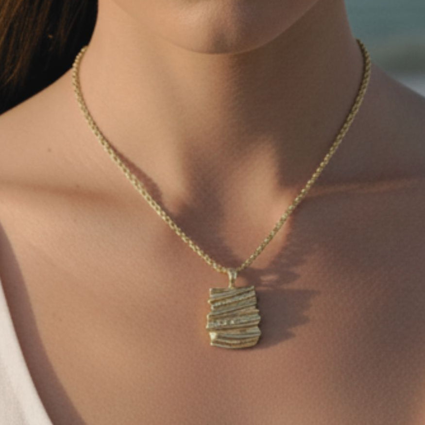 Close-up of a woman wearing a gold necklace with a textured pendant against a blurred natural background.