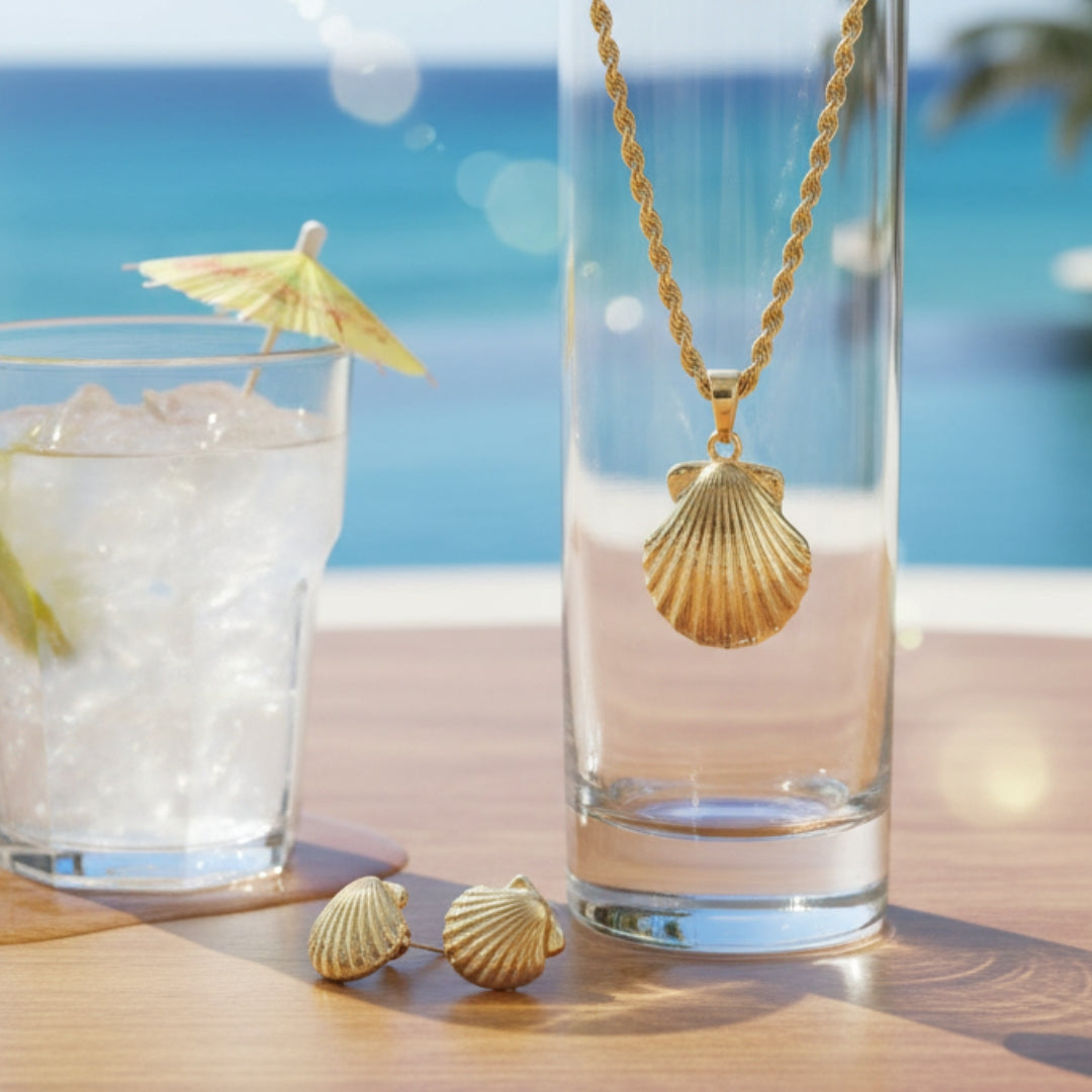 Gold shell necklace and earrings on a beach setting with a glass of ice.