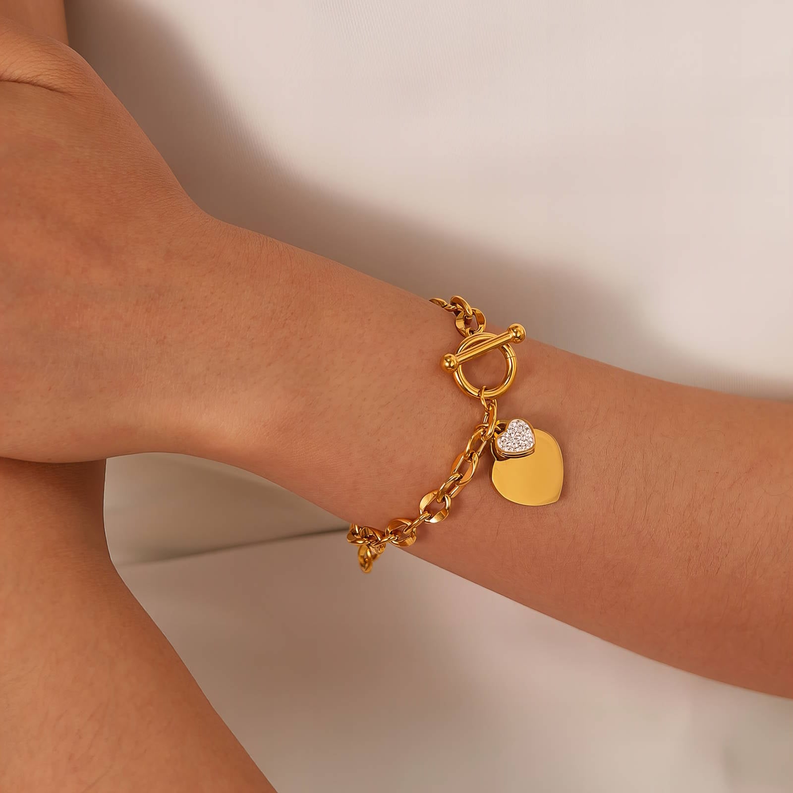 Gold bracelet with heart and round tag charm on a wrist against a neutral background